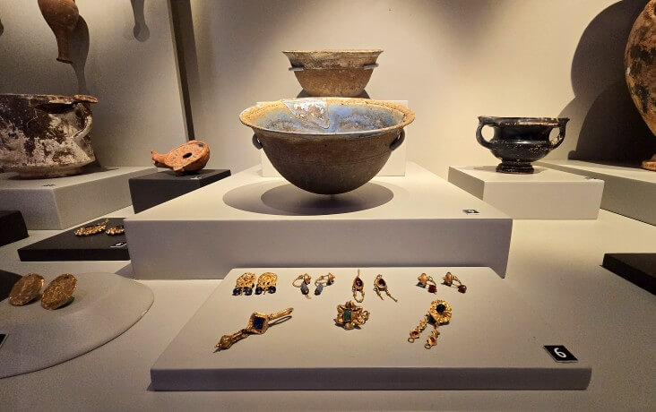 İznik Müzesi’nde sergilenen Hellenistik döneme ait cam kaplar, metal kaplar ve değerli taşlarla süslenmiş altın takı örneklerinin vitrindeki detaylı düzenlemesi.